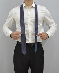 A man wearing a white long-sleeve dress shirt and black trousers models a dark blue patterned necktie. He is pointing at the tie with both hands against a plain gray studio background.