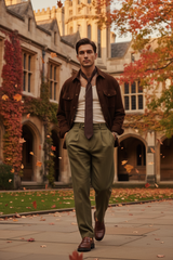 A man walking outdoors in front of a historic stone building with autumn foliage. He is wearing a brown jacket, white shirt, dark tie, olive green trousers, and brown shoes.
