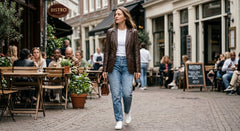 A woman walking down a cobblestone street wearing a brown leather jacket, white t-shirt, blue jeans, and white sneakers, with outdoor cafe seating and restaurant storefronts in the background.