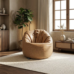 A tan faux-leather bean bag armchair with a slouchy, oversized design, styled in a warm, modern living room setting with a textured rug, wooden furniture, and a potted fiddle-leaf fig plant.