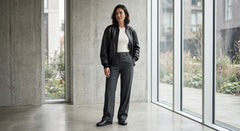 A woman stands in a minimalist concrete building interior wearing a black leather jacket, white top, and dark gray wide-leg trousers.