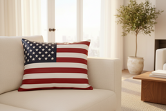 A square throw pillow featuring the American flag design, resting on a cream-colored sofa in a bright, modern living room.