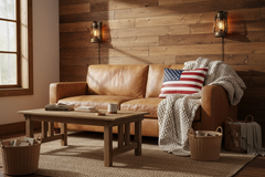 A warm rustic living room scene featuring a tan leather sofa with a red, white, and blue American flag throw pillow and a cream knit blanket draped over the arm. A matching wooden coffee table sits in front on a beige rug, with a wood plank wall, wall sconces, and a window in the background.