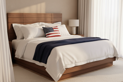 A modern wooden bed frame dressed in white bedding with a navy blue throw blanket and a small American flag accent pillow, set in a softly lit bedroom with curtains and a bedside lamp.
