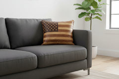 A square decorative throw pillow with an American flag design made from brown and tan leather, resting on a brown leather armchair in a cozy room setting.