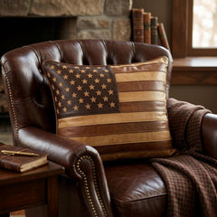 A decorative square throw pillow featuring an American flag design in shades of brown and tan, styled on a dark brown leather armchair in a cozy room setting.