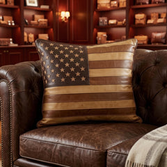 A square decorative throw pillow featuring a rustic American flag design made from brown and tan leather, resting on a dark brown leather tufted armchair in a cozy living room setting.