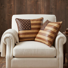 Two decorative throw pillows with a rustic American flag design made from wood or faux leather stripes and stars, styled on a cream upholstered armchair with a knitted throw blanket.