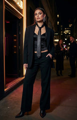 A woman stands on a city sidewalk at night wearing a black tailored outfit with a structured blazer, wide-leg trousers, a black satin-like top, a patterned black tie, and black pointed heels. Storefront lights and blurred pedestrians are visible in the background.