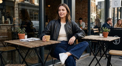 A woman sits at an outdoor cafe table wearing a black leather jacket, cream top, blue jeans, and white sneakers, holding a coffee cup.