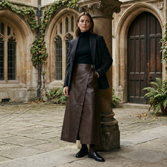 A woman stands in a stone courtyard wearing a dark charcoal blazer, black turtleneck, brown leather wrap skirt, and black loafers, leaning against a decorative column.