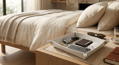 A white rectangular tray with small gold-toned corner accents sits on a wooden bedside table in a softly lit bedroom. The tray holds a watch, wallet, sunglasses, and a phone, with a bed and pillows in the background.