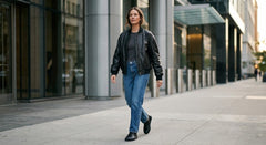 A woman walking on a city sidewalk wearing a black leather bomber jacket, black t-shirt, blue jeans, and black shoes.