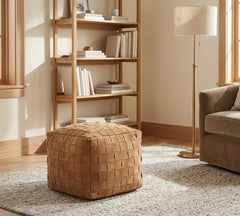 A square woven leather pouf ottoman in a warm tan-brown color, styled in a cozy living room setting on a gray rug next to a wooden bookshelf and floor lamp.