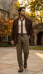 A man walking outdoors on a stone path in front of a historic stone building with autumn trees. He is wearing a brown suede jacket, a cream shirt, a dark tie, olive green trousers, and brown leather shoes.