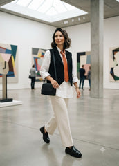 A woman walks through a modern art gallery wearing a stylish outfit consisting of a white button-down shirt, a black vest, an orange tie, cream wide-leg trousers, and black loafers, while carrying a black shoulder bag.