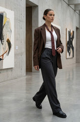 A woman walks through a modern art gallery wearing a brown blazer, white top, burgundy tie, and dark charcoal trousers.