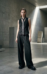 A model stands in a minimalist concrete gallery space wearing a black sleeveless vest, wide-leg black trousers, a patterned black-and-white necktie, and black platform loafers.