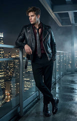 A man wearing a black leather moto jacket, black trousers, and a dark patterned tie stands on a rooftop balcony at night with a city skyline in the background.