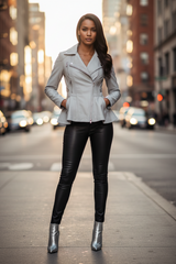 A woman stands on a city street at sunset wearing a light gray moto-style peplum jacket, black faux-leather pants, and silver ankle boots.