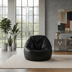 A black leather bean bag chair centered on a light beige rug in a modern, sunlit living room with large windows, plants, and a sideboard.