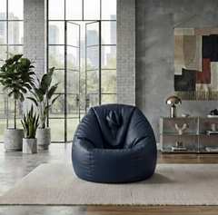 A dark navy blue bean bag chair placed on a light beige rug in a modern loft-style room with large industrial windows, brick walls, potted plants, and a side table.