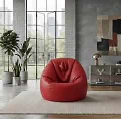 A red faux-leather bean bag armchair is placed in a modern, industrial-style living room with a concrete wall, large windows, and potted plants.