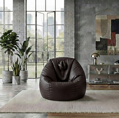 A dark brown leather bean bag chair placed on a light beige rug in a modern industrial loft room with large windows, brick walls, and plants.