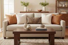 A light beige upholstered loveseat sofa in a styled living room, decorated with five throw pillows in cream, tan, and brown, set behind a dark wood coffee table.