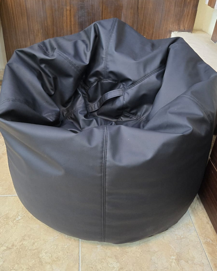 A large black bean bag chair made of durable fabric, placed on a beige tiled floor in front of a dark wood-paneled wall.