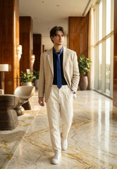 A man walking down a luxury hotel lobby wearing a beige linen-blend suit, featuring a beige blazer, cream trousers, a navy blue shirt, a bright blue tie, and white sneakers.