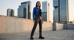 A woman stands on a rooftop wearing a blue leather jacket, black top, black leather pants, and black boots, with a city skyline in the background.