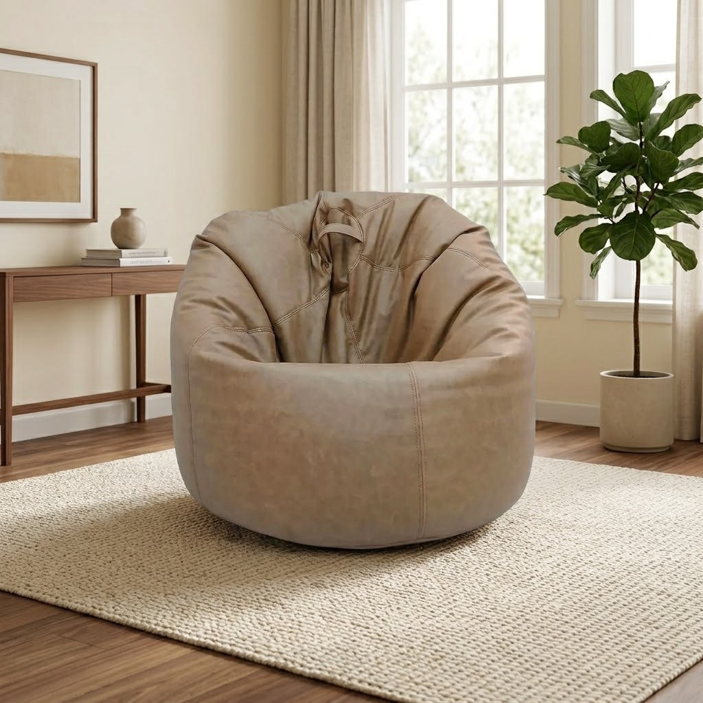 A round, oversized lounge chair in a taupe-beige suede-like fabric, styled in a bright, modern living room with a woven rug, wooden console table, and a potted fiddle-leaf fig plant.