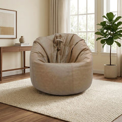 A round, oversized lounge chair in a taupe-beige suede-like fabric, styled in a bright, modern living room with a woven rug, wooden console table, and a potted fiddle-leaf fig plant.