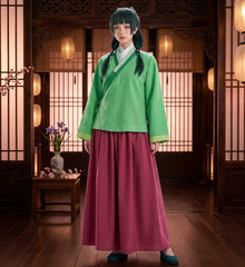 A person wearing a traditional-style outfit with a bright green long-sleeve top and a long magenta skirt, standing in a wooden interior with lanterns and screens.