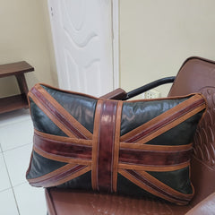 A rectangular black leather travel pillow featuring a Union Jack flag design with brown leather straps, resting on a brown leather chair in an indoor room.