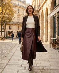 A woman walking down a cobblestone street wearing a dark brown long coat, a cream ribbed top, and a dark brown faux-leather wrap skirt, carrying a brown handbag.