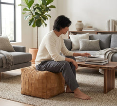 A man sits barefoot on a woven tan pouf in a bright, modern living room, reading books on a wooden coffee table next to a gray sofa.