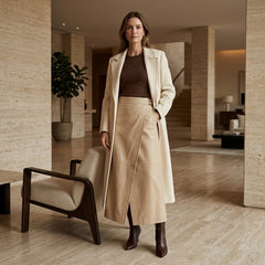 A woman stands in a modern indoor setting wearing a long cream coat over a brown top and a beige midi skirt, paired with dark brown ankle boots.