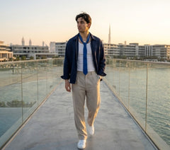 A man walking on a glass-railled walkway at sunset, wearing a navy blue overshirt, a white shirt, a blue tie, beige trousers, and white sneakers, with a city skyline in the background.