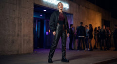 A woman stands on a city sidewalk at night wearing a black bomber jacket, black leather pants, and a dark burgundy top. The background shows a concrete building entrance with blurred people and a lit sign.