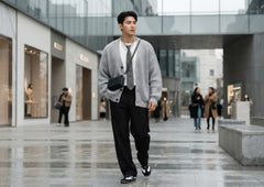 A man walking in a modern outdoor shopping district, wearing a light gray knit cardigan over a white shirt and dark tie, paired with black trousers and black-and-white sneakers.