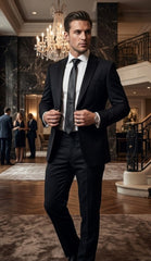 A man wearing a black formal suit, white dress shirt, and dark tie standing in a luxurious indoor lobby with marble walls, a chandelier, and a staircase in the background.
