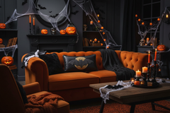 A Halloween-themed living room scene featuring a tufted orange sofa with black throw blankets and a Batman-patterned pillow, surrounded by spooky decor including cobwebs, candles, jack-o'-lanterns, and a dark fireplace.