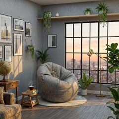 A cozy modern living room corner featuring a large gray bean bag chair on a round rug, positioned in front of a large black-framed window with a city skyline at sunset. The room has gray walls decorated with framed art, a wooden shelf with hanging plants, and a gold floor lamp.
