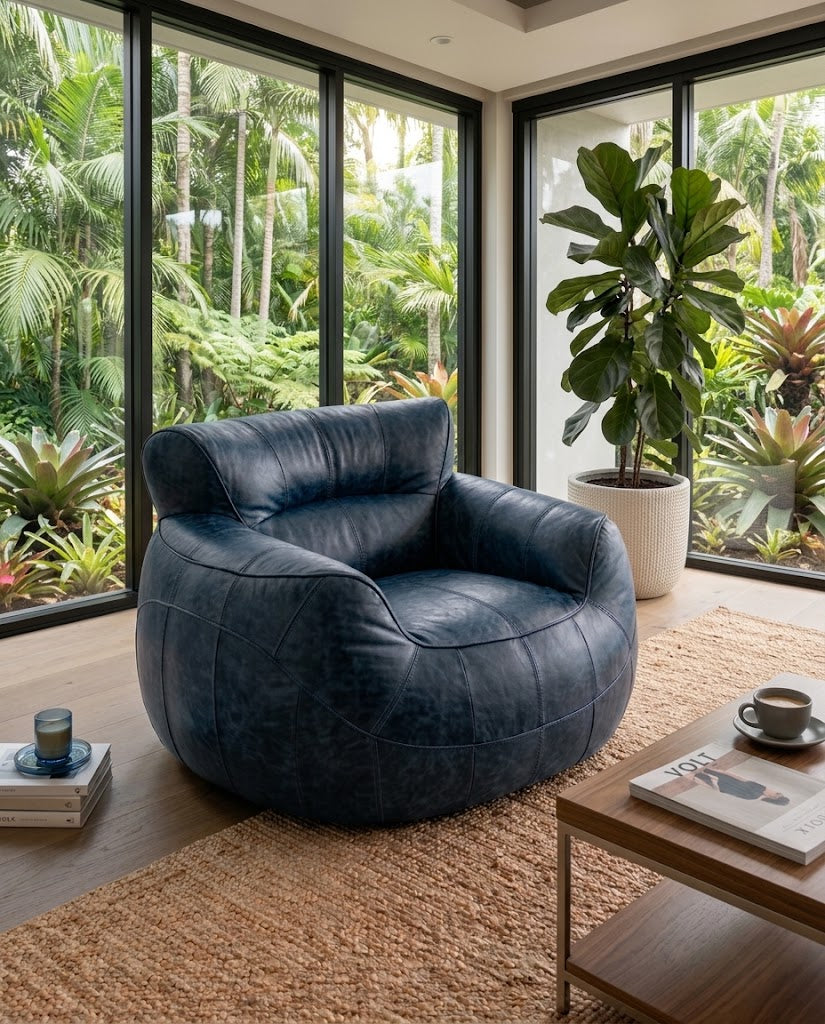 A dark navy blue leather club chair in a modern living room setting with large windows overlooking a lush green garden, a woven rug, and a wooden coffee table.