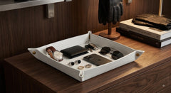 A white leather organizer tray with brass corner hardware, styled on a dark wood dresser holding a watch, wallet, sunglasses, earrings, and a smartphone. Books and a black glove stand are visible in the background.