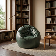 A dark green leather bean bag chair in a cozy, rustic library setting with wooden bookshelves, a floor lamp, and a small wooden side table.