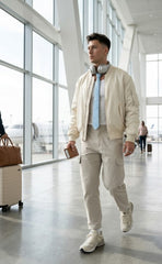 A man walking through an airport terminal wearing a cream-colored bomber jacket, matching cargo pants, a light blue tie, and white sneakers, with headphones around his neck.