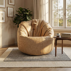 A round, oversized tan leather armchair with a slouchy, beanbag-like silhouette, placed in a sunlit living room next to a small wooden side table with books and a mug.